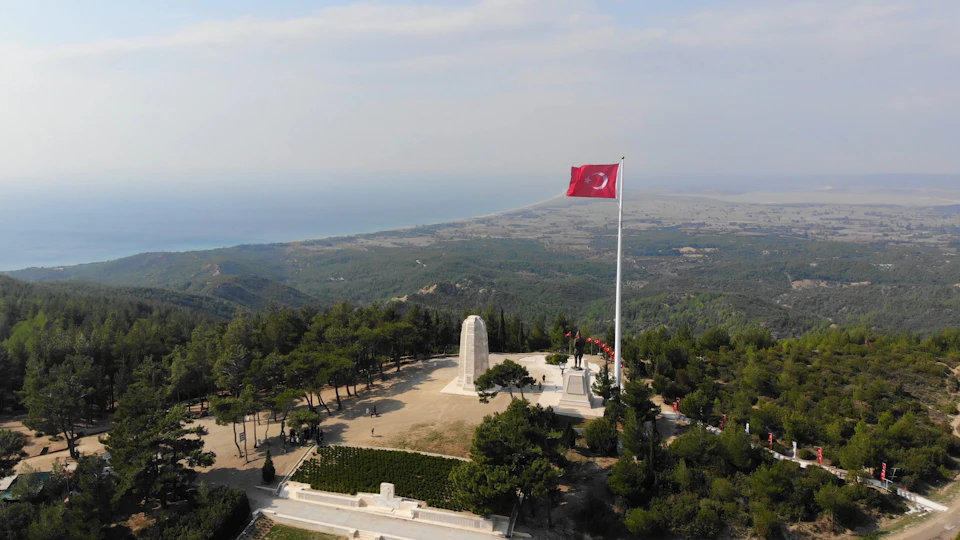 Aerial view of a monument with a large flagpole flying a red flag, surrounded by trees and overlooking a distant coastline.
