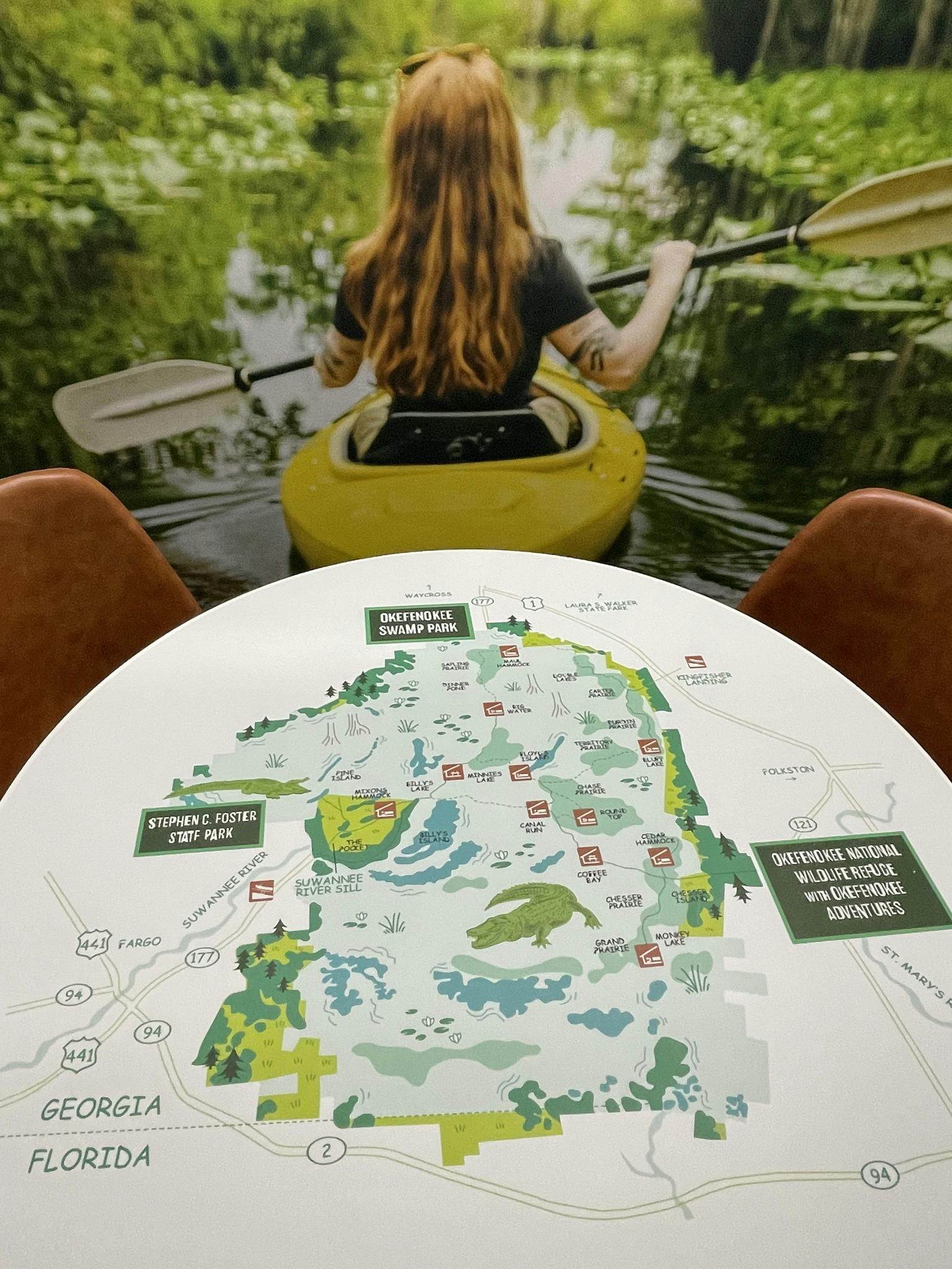 Kvinde i kajak på vand, set bagfra. Nedenfor er et kort over Okefenokee Swamp Park på et bord med tekst og illustrationer.
