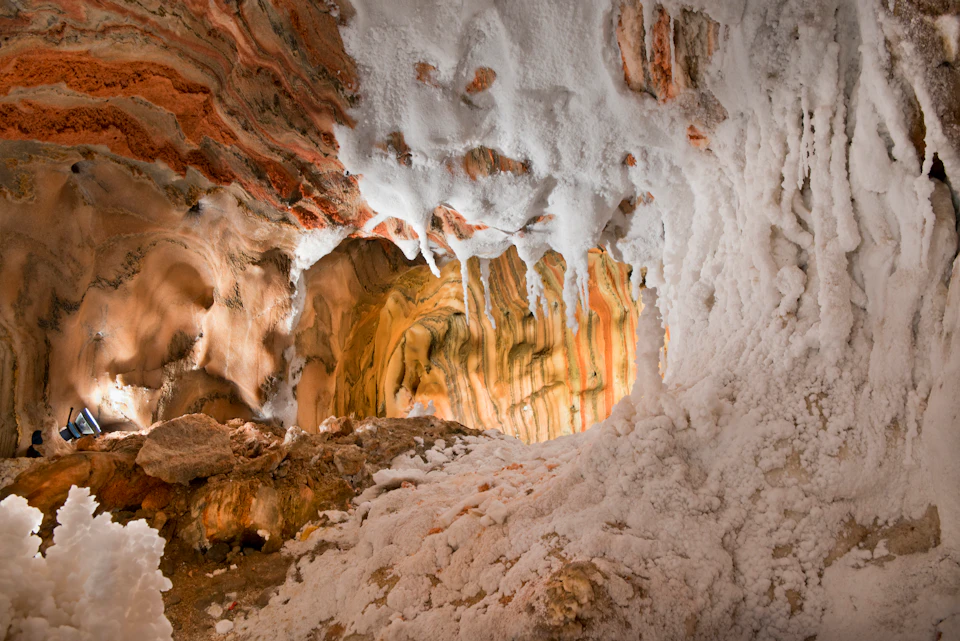 A cave interior with orange and white stalactites and stalagmites, featuring a textured, layered ceiling and walls.