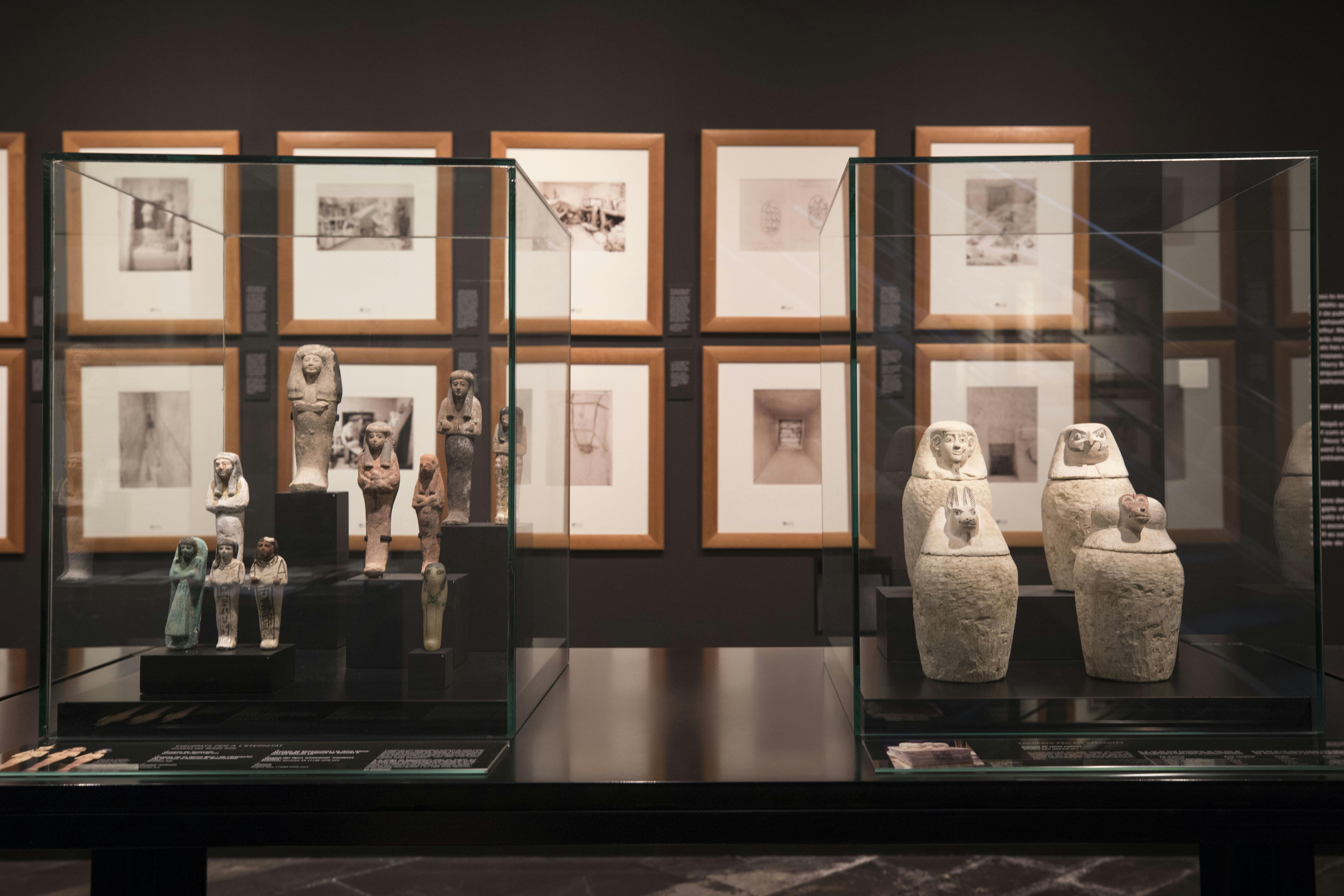 Ancient artifacts displayed in glass cases, with framed photographs on the wall behind them in a museum setting.