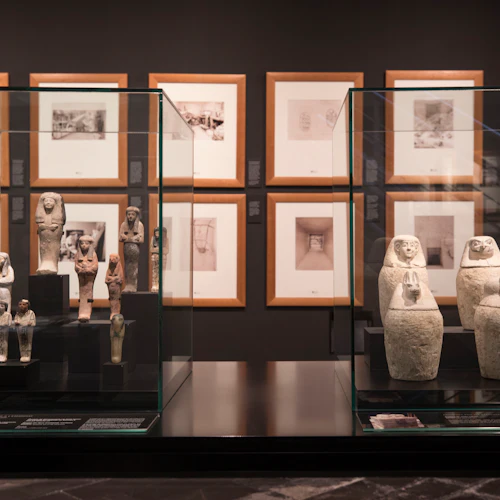 Forntida artefakter visas i glasfodral, med inramade fotografier på väggen bakom dem i en museumsmiljö.