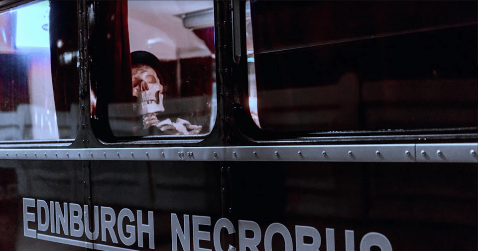 A skeleton wearing a hat is seated inside a dark bus, visible through a window with partially obscured lettering on the exterior.