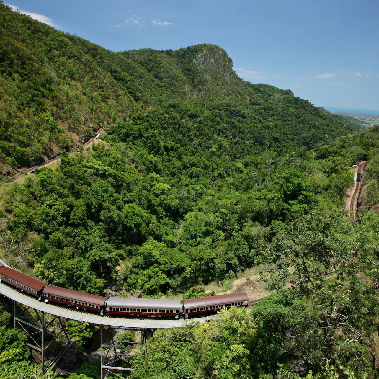 A train navigates a curved track through a lush, green mountainous landscape under a clear blue sky.