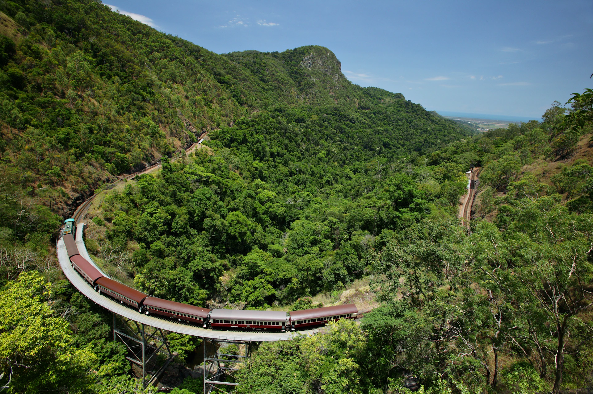 A train navigates a curved track through a lush, green mountainous landscape under a clear blue sky.