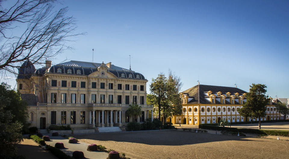 Two historic buildings with ornate architecture stand facing an open courtyard, surrounded by trees under a clear blue sky.