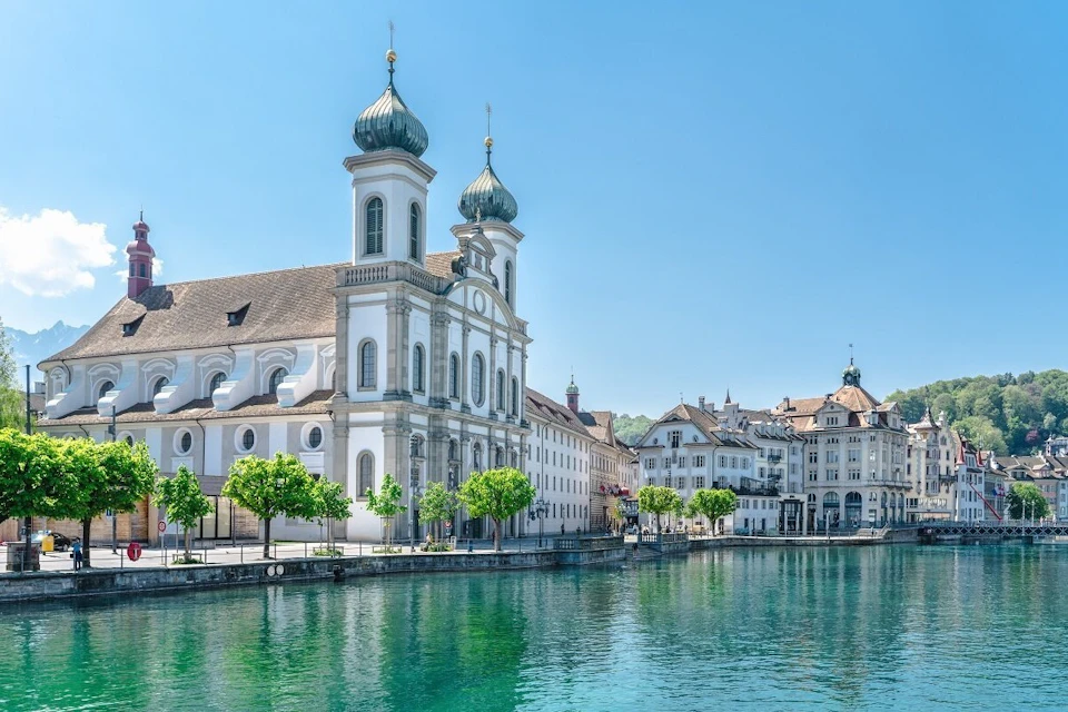 A waterfront scene with a historic building featuring twin green domes, trees, and clear water under a blue sky.