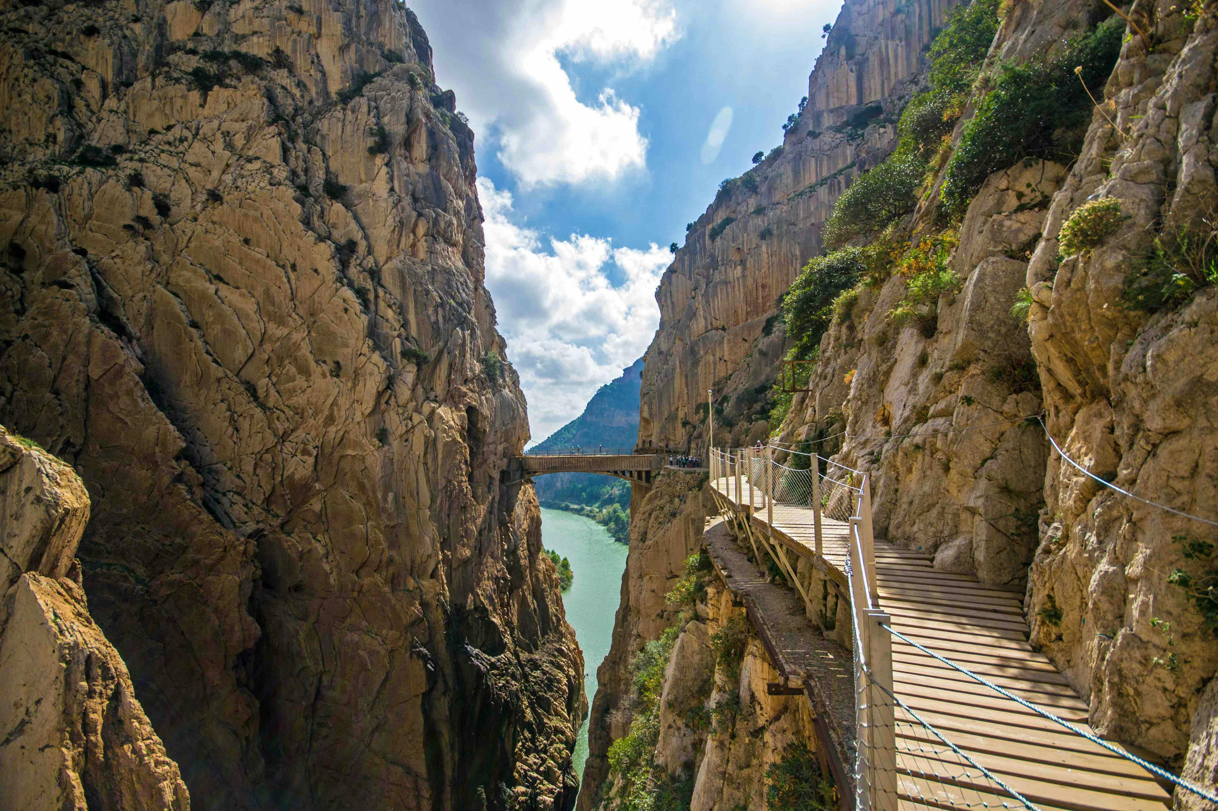 A narrow gorge with a hanging bridge and a wooden walkway along steep rocky cliffs under a bright, partly cloudy sky.