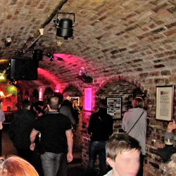 Trångt tegelväggigt rum med välvt tak, människor som tittar på en solomusiker som uppträder på scenen, färgglada lampor ovanför.