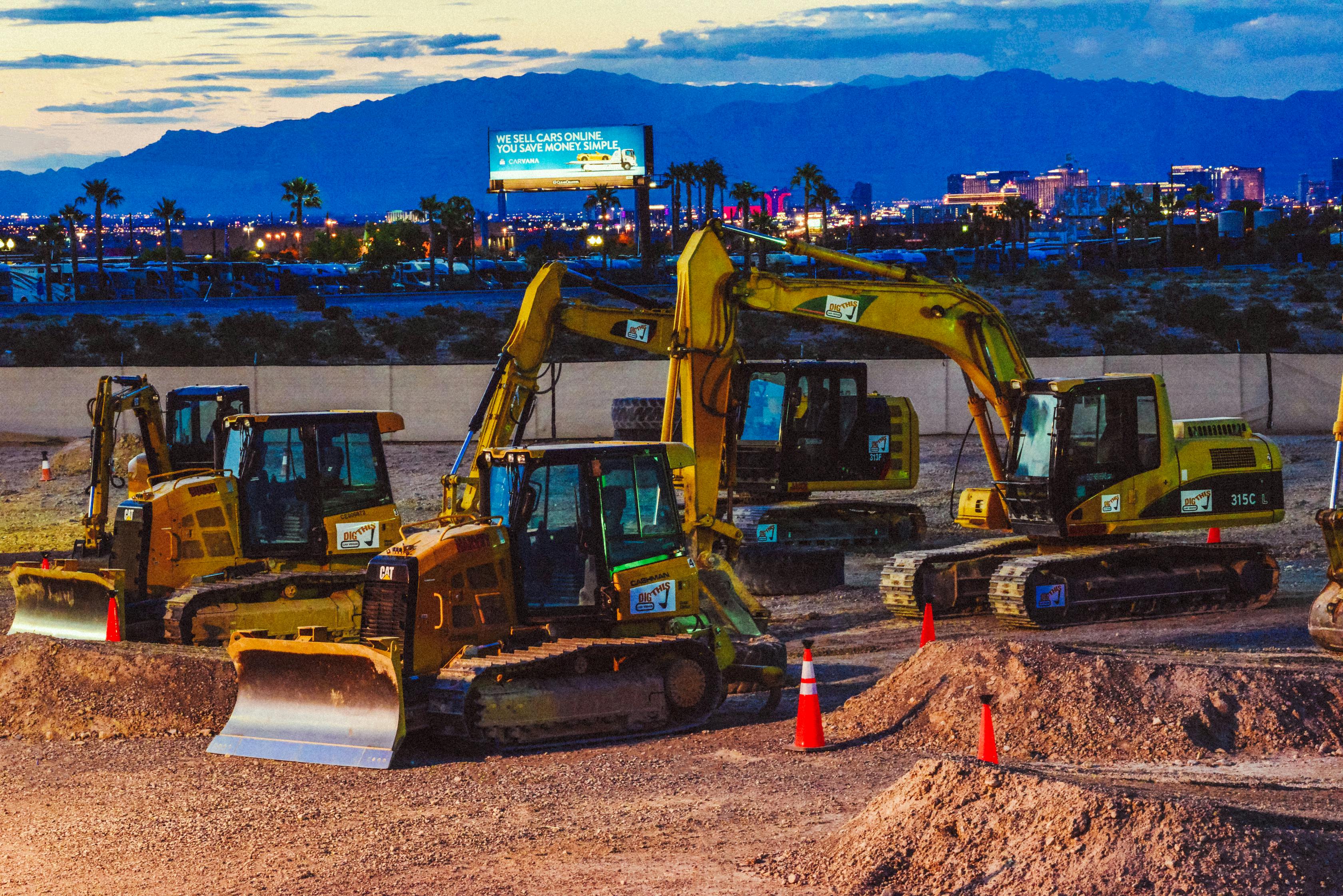 해질 무렵 교통 콘, 도시의 불빛, 산을 배경으로 건설 현장에 노란색 굴삭기 4대가 주차되어 있습니다.