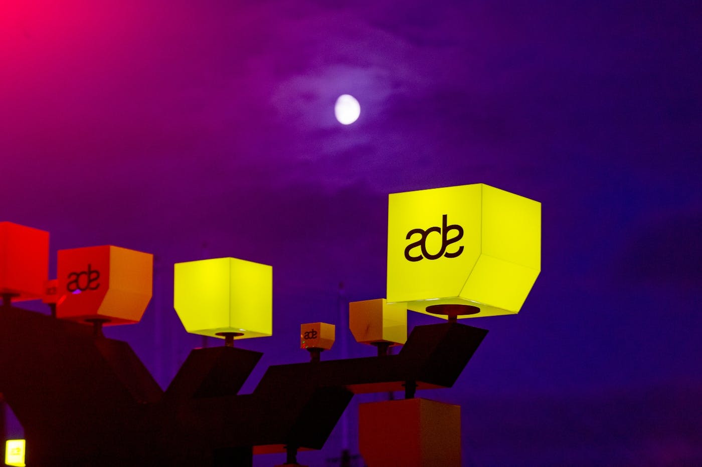 Osvětlené kostky s „ade“ logy na soumračné obloze se zářícím měsícem v pozadí.