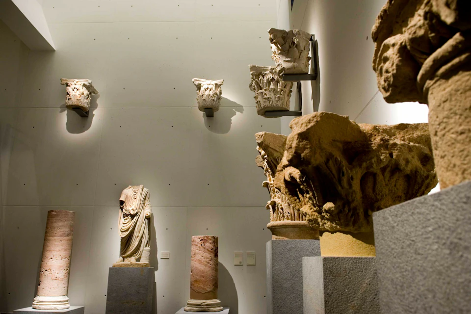 Museum exhibit with ancient sculptures, including a headless statue, decorative capitals, and a stone column, all on display stands.