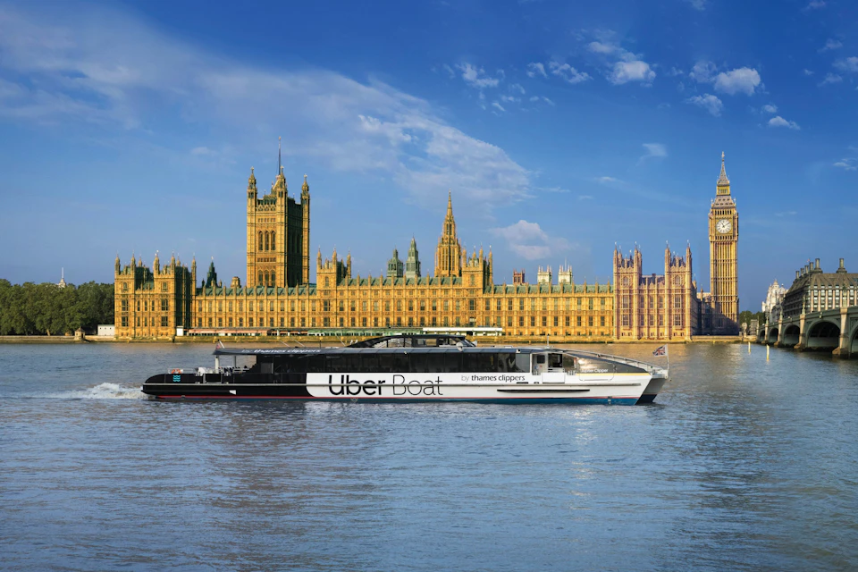 An Uber Boat by Thames Clippers ferry on the Thames River in front of the Palace of Westminster under a blue sky.