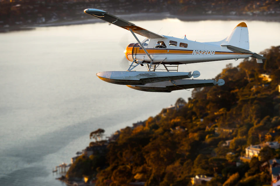 A small white and yellow seaplane flying over a coastal landscape with water and shoreline below.