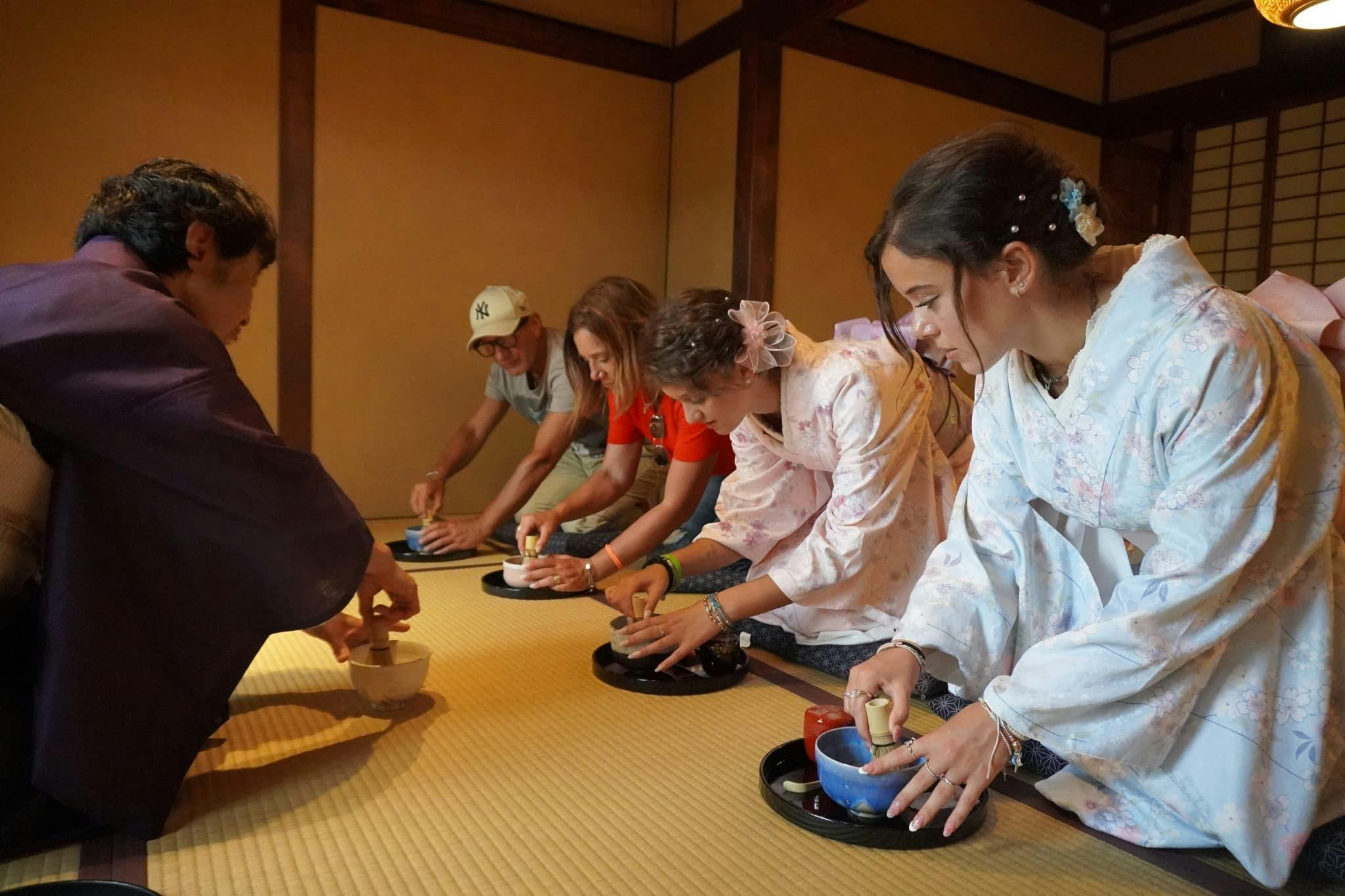 A group of people participate in a traditional tea ceremony, preparing tea on a tatami floor in a Japanese-style room.