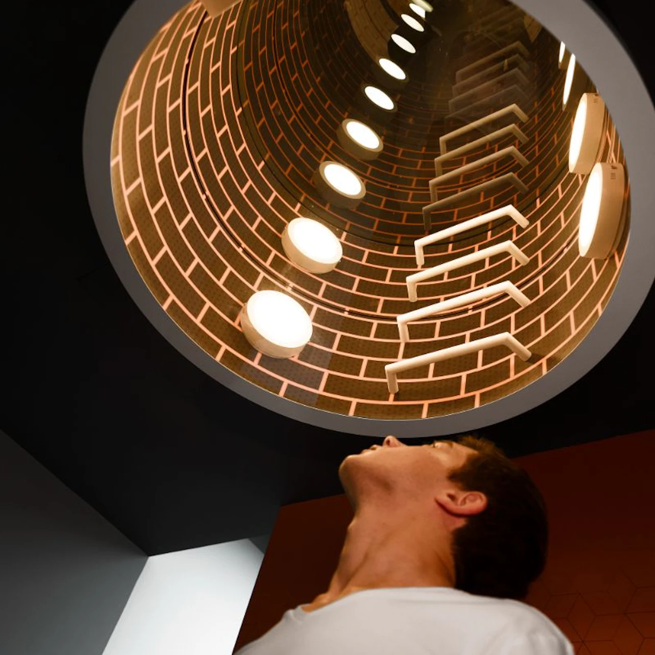 Person looking up at a circular, brick-patterned tunnel with illuminated round lights and ladder-like structures.