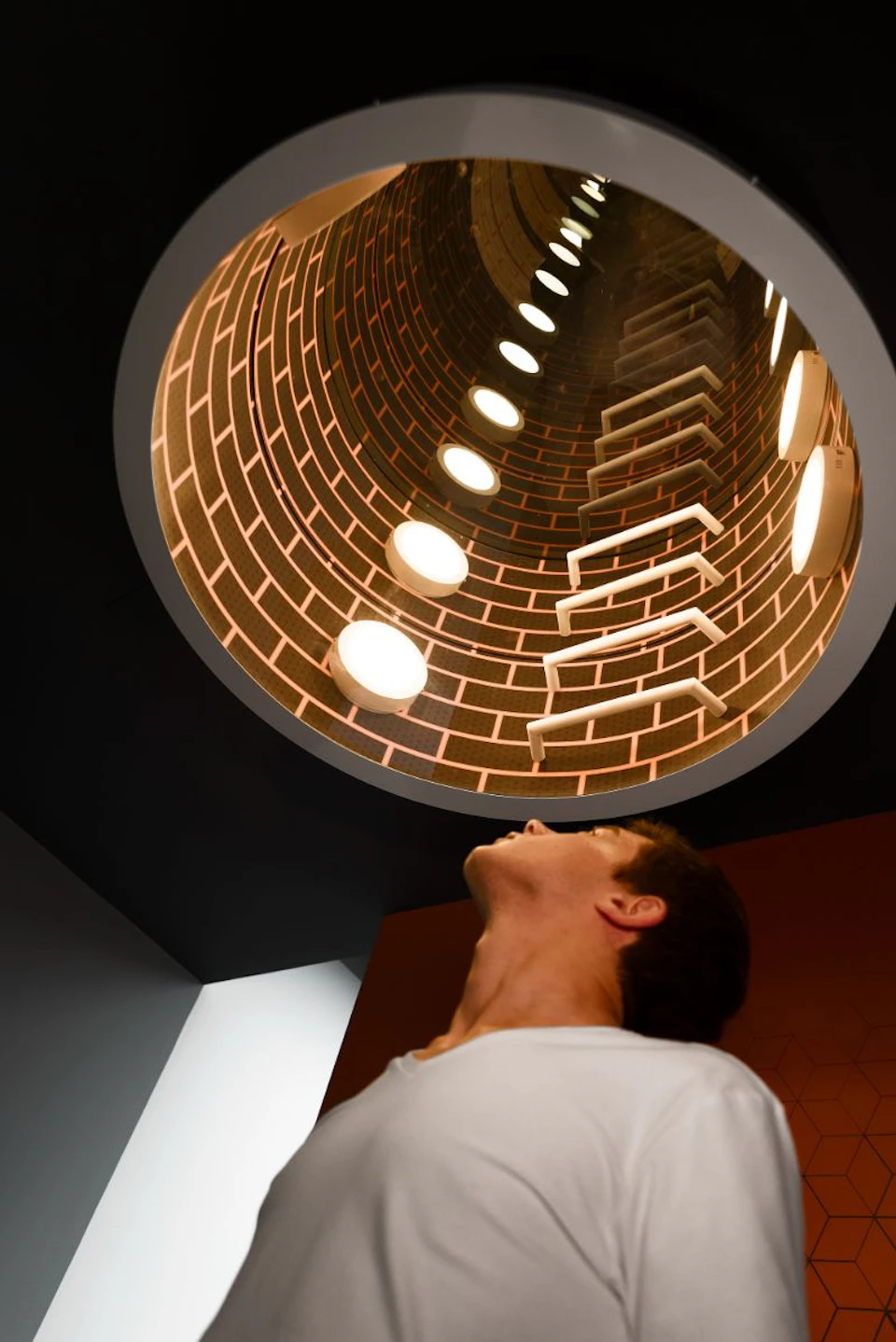 Person looking up at a circular, brick-patterned tunnel with illuminated round lights and ladder-like structures.