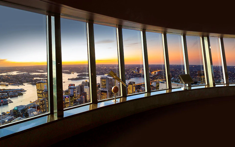 City skyline and harbor view at sunset seen through tall windows, with a telescope on an observation deck in the foreground.