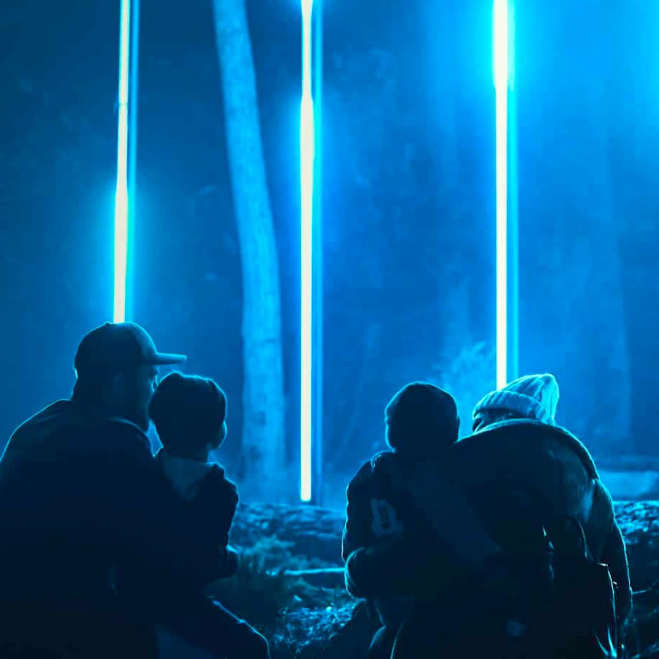 Silhouetted people sitting under blue glowing vertical lights in a forested area at night.
