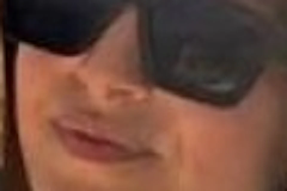 A person wearing a hat and sunglasses is on a boat near rocky cliffs surrounded by a blue sea.