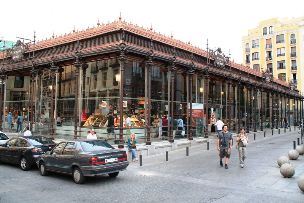 Personnes marchant près du Mercado de San Miguel, un marché historique avec des murs en verre, des étals intérieurs visibles et des voitures garées à l'extérieur.