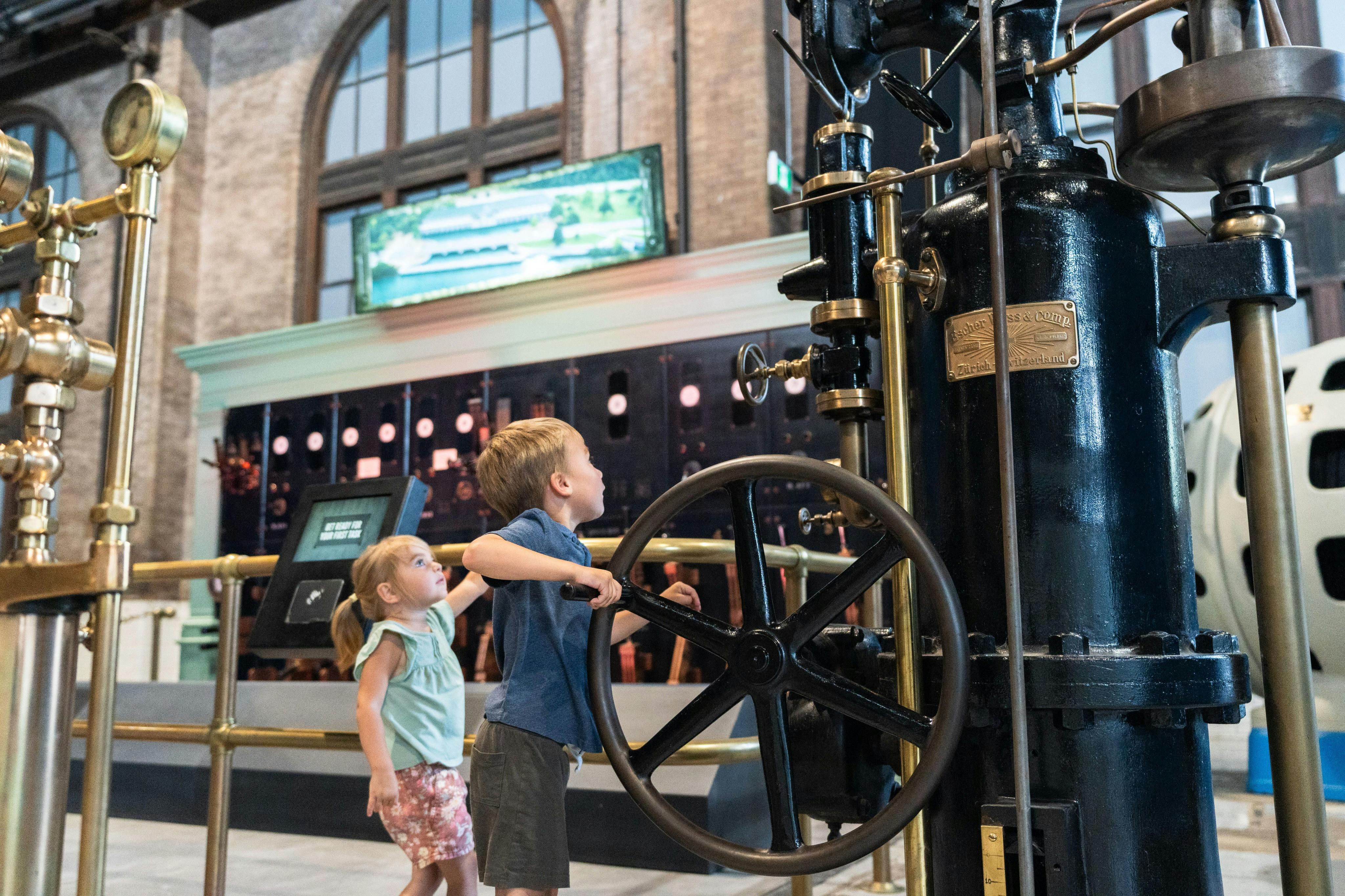 두 명의 어린이가 산업적이고 역사적인 장식이 있는 박물관에서 커다란 기계 바퀴 전시물과 상호 작용합니다.