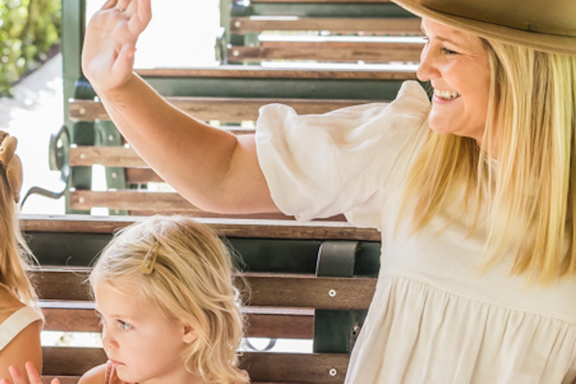 A woman and three young girls wave while sitting on a green open-air tram in a lush, sunny environment.