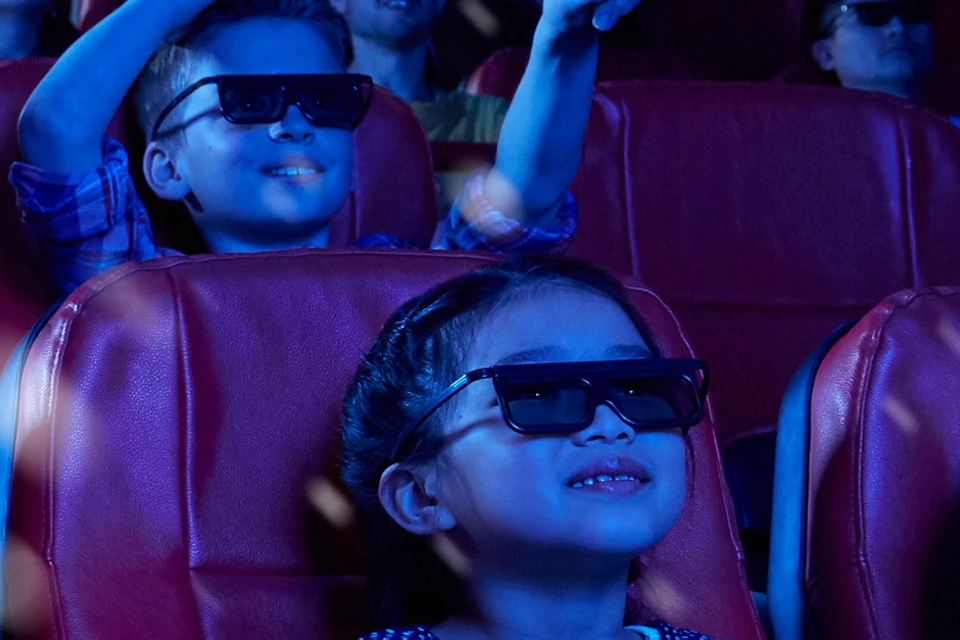 People wearing 3D glasses sit in a theater with red seats, watching a film amidst floating particles, with children in the front row.