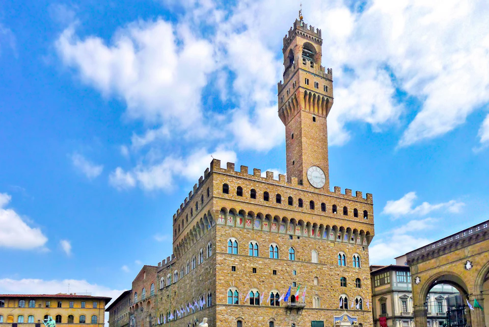Medieval-style building with a tall clock tower under a partly cloudy sky. Flags hang from the middle row of arched windows.
