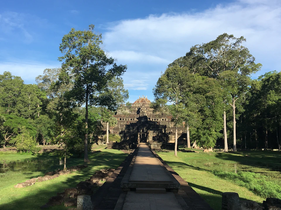 Ancient stone temple surrounded by tall trees and lush greenery under a blue sky with a paved pathway leading to the entrance.