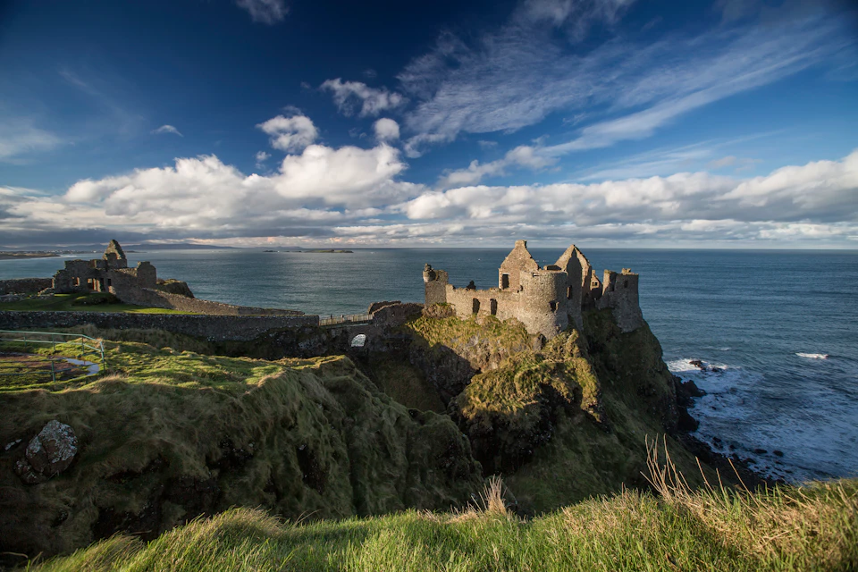 Ancient stone castle ruins sit on a cliff overlooking a vast, calm ocean under a partly cloudy blue sky.