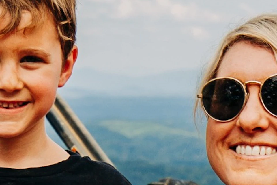A woman and a boy stand smiling next to an off-road vehicle on a scenic hilltop with a view of forested mountains.