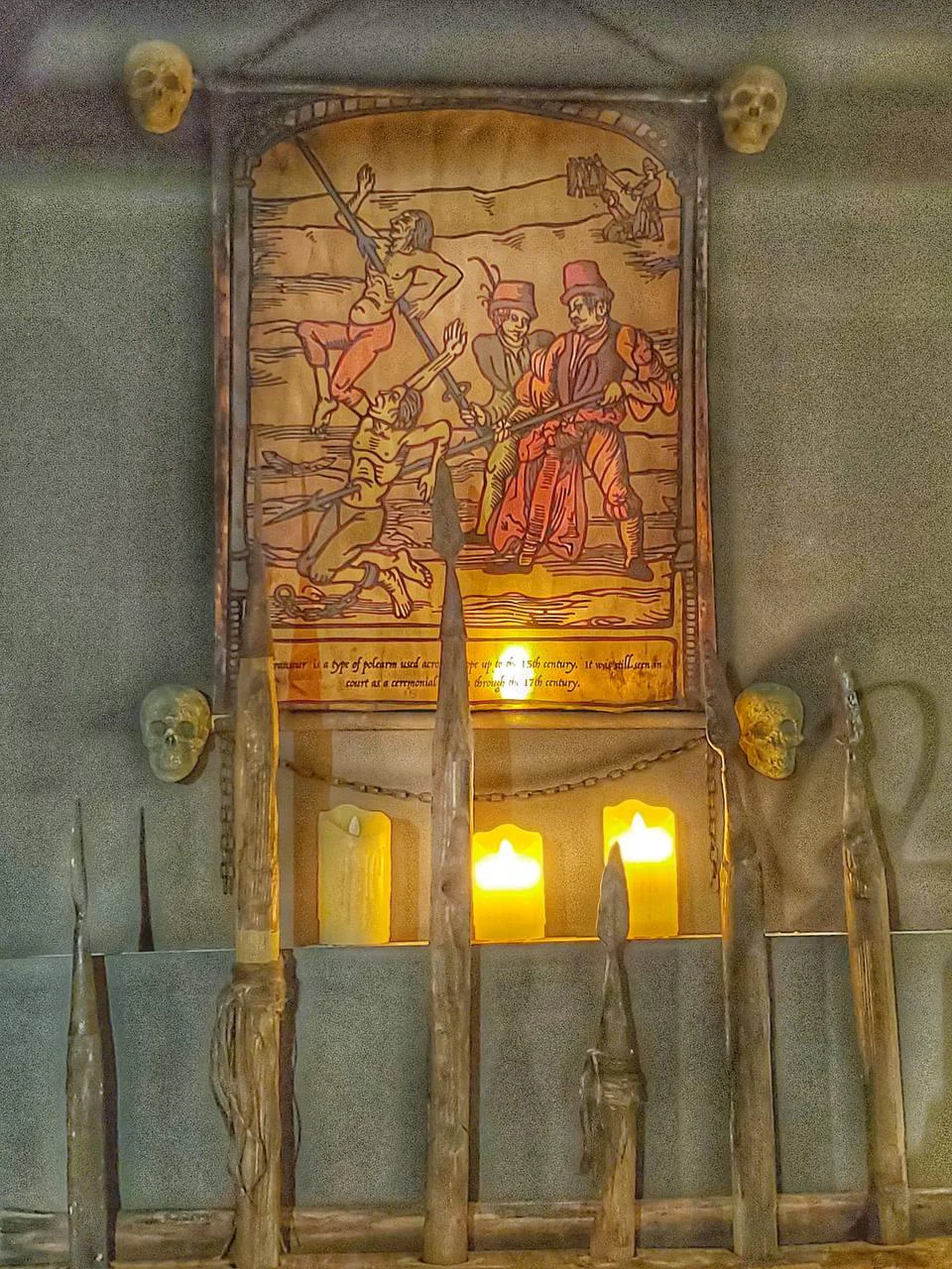 An old illustration depicting a battle scene, with skulls, spears, and lit candles placed below it.