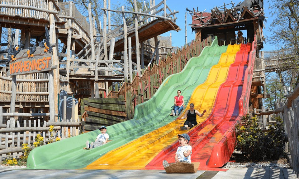 Children and adults sliding down colorful slides at a playground with a wooden structure in the background.