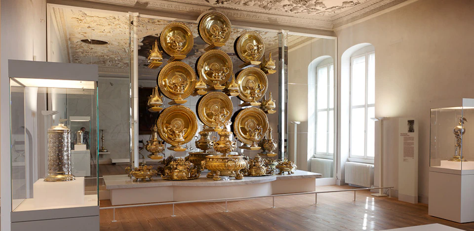 A display of ornate gold dishes and vessels on shelves in a decorated room with a wooden floor and arched windows.