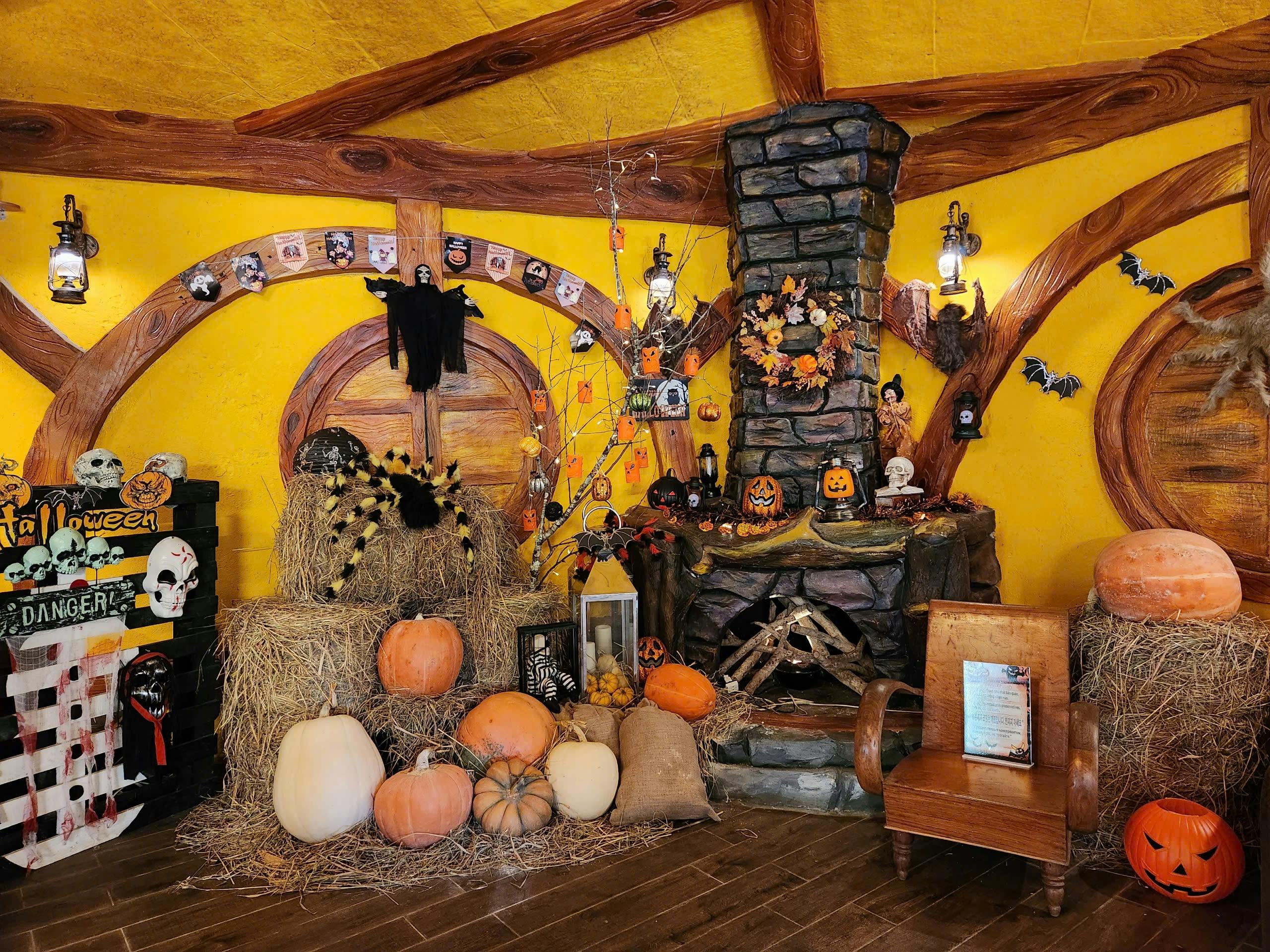 A cozy Halloween-themed room with pumpkins, hay bales, spooky decorations, and a stone fireplace on a wooden floor.