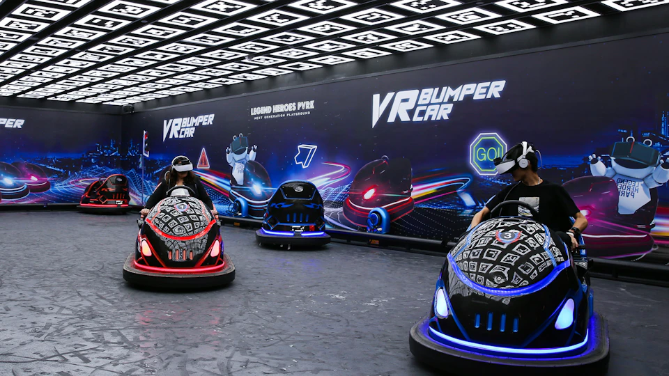 People using virtual reality headsets in illuminated bumper cars on an indoor track with futuristic branding on the walls.