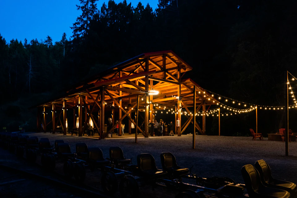 Outdoor wooden pavilion at dusk, adorned with string lights, and several people gathered underneath. Forest backdrop.