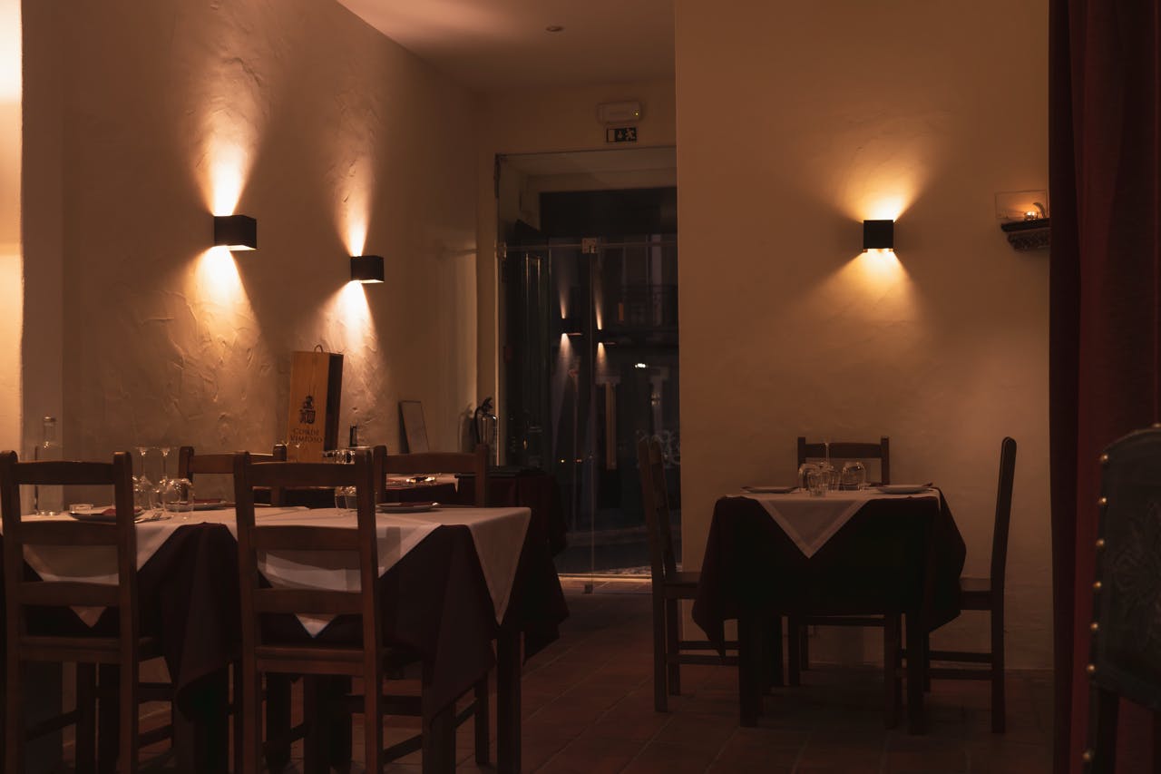 木製のテーブルと椅子、白いテーブルクロス、壁に取り付けられた照明が暖かい雰囲気を作り出す薄暗いレストラン。