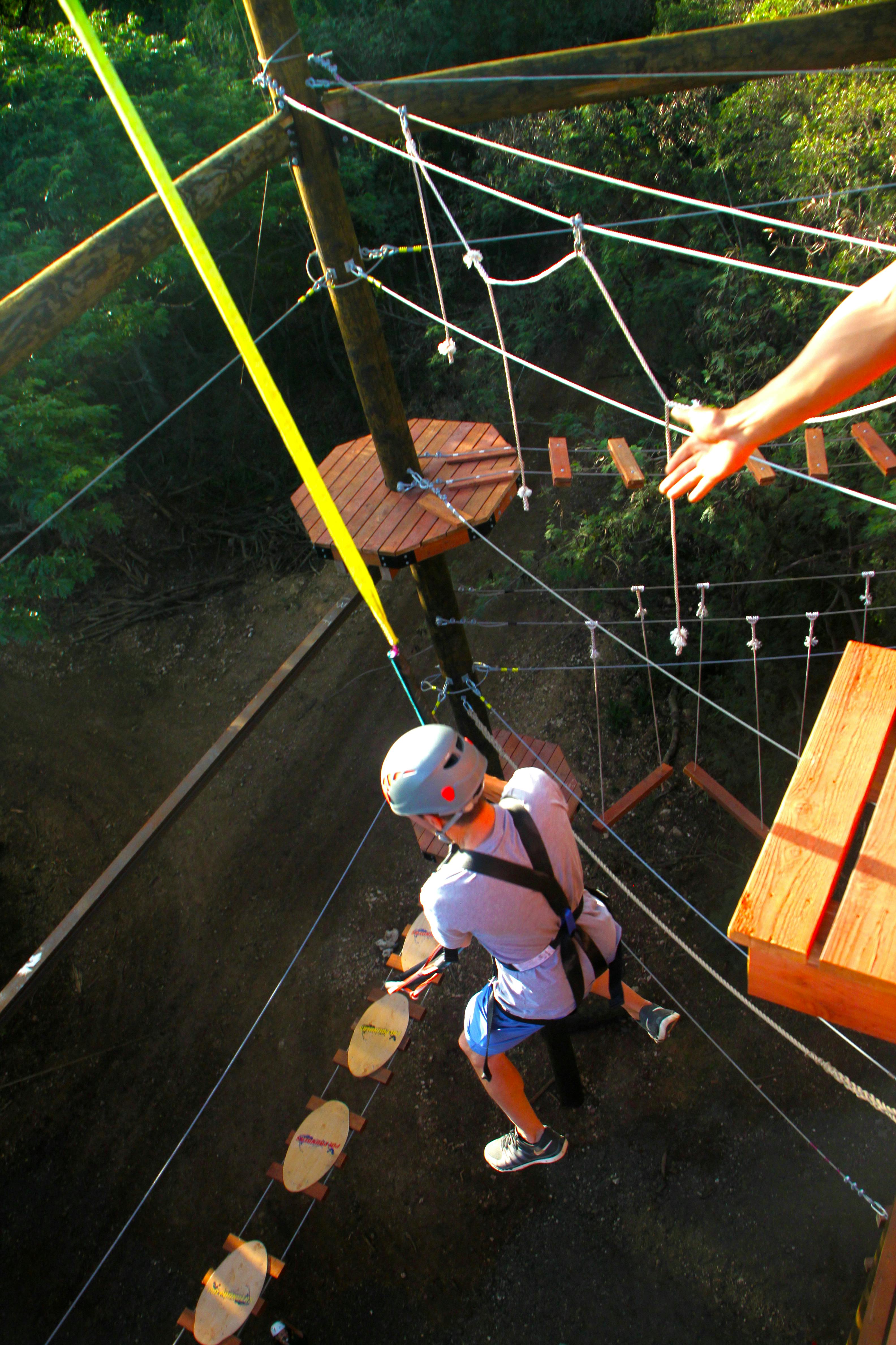 헬멧과 하네스를 착용한 사람이 나무와 나무 플랫폼으로 둘러싸인 지상 높이의 로프 코스를 항해합니다.