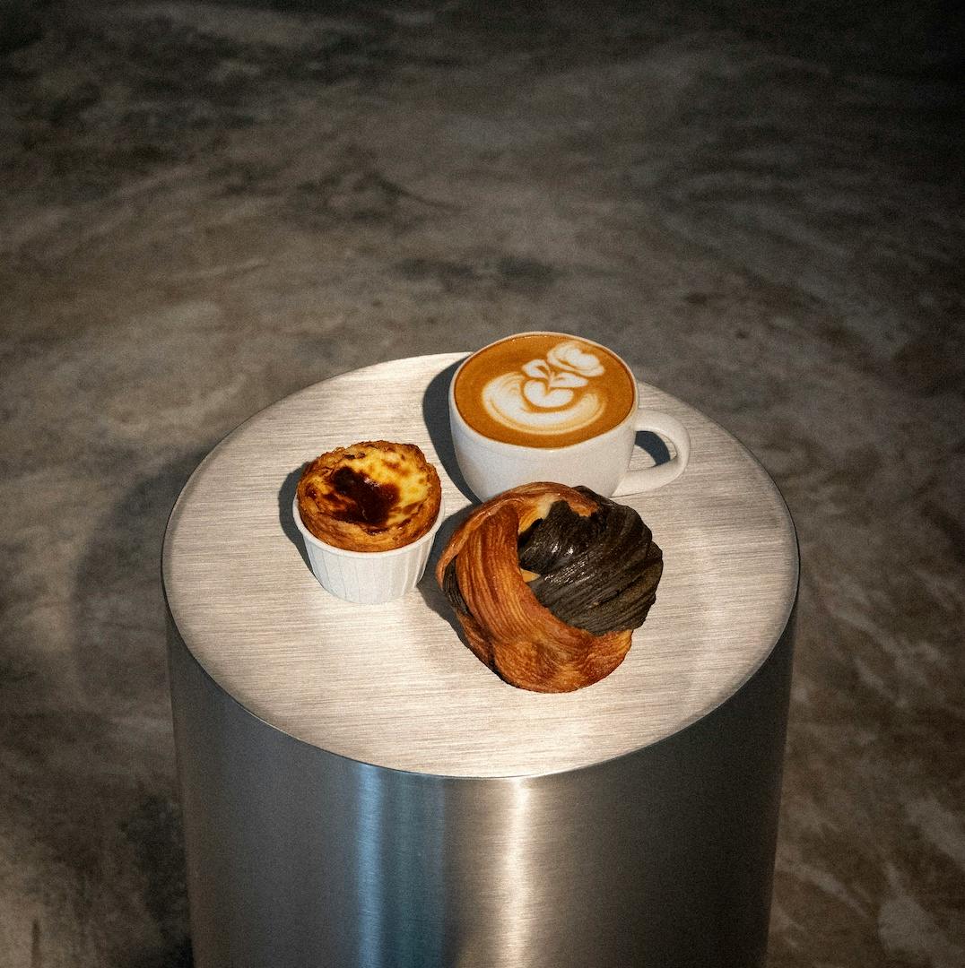 A latte with latte art, a custard tart, and a twisted pastry on a round metal table, set against a concrete floor.