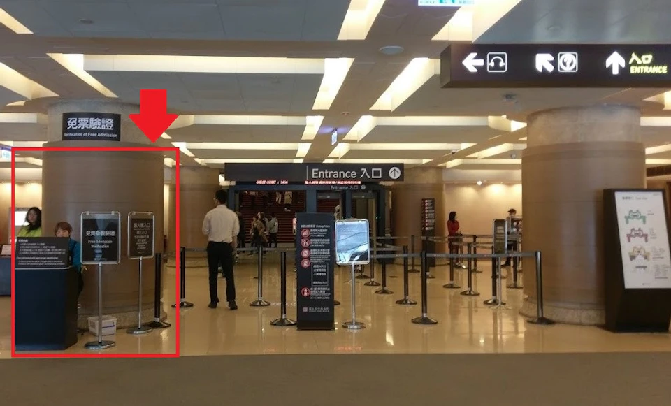 A queue area with an 'Entrance' sign, barriers for lines, and several people walking or standing in the background.