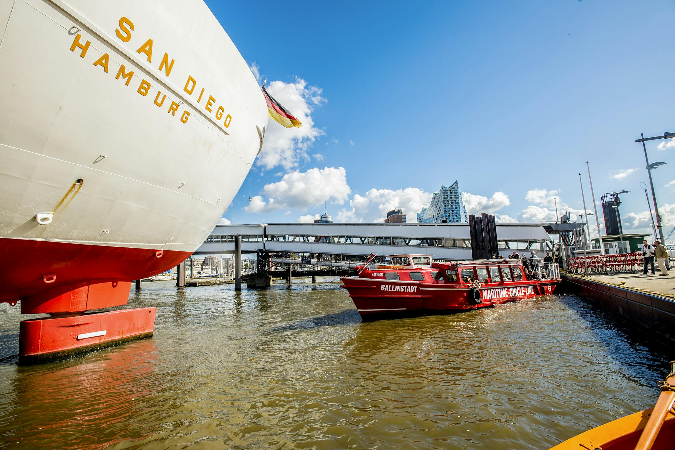 Un grand navire portant l'inscription "San Diego" et un bateau touristique rouge avec des personnes à bord dans un port sous un ciel ensoleillé avec des nuages épars.