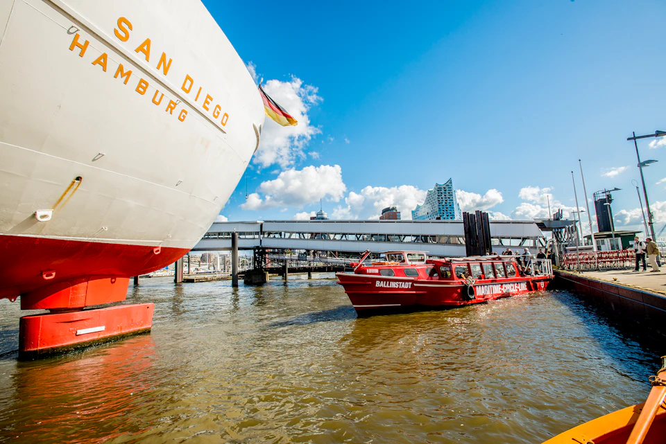 A large ship labeled "San Diego" and a red tour boat with people on board in a harbor under a sunny sky with scattered clouds.