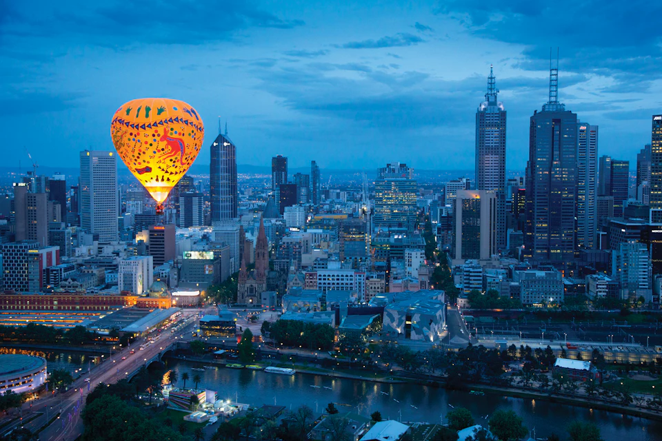 A colorful hot air balloon floats above a city skyline with tall buildings and a river at dusk.