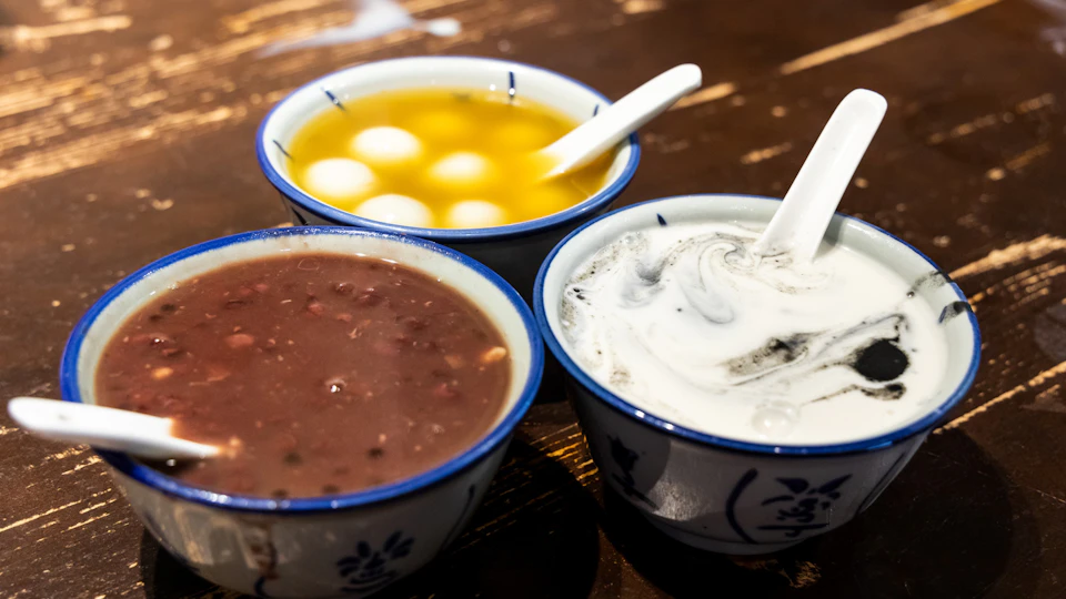 Three bowls of dessert; one with yellow broth and white balls, one with red bean soup, and one with a creamy, black sesame swirl.