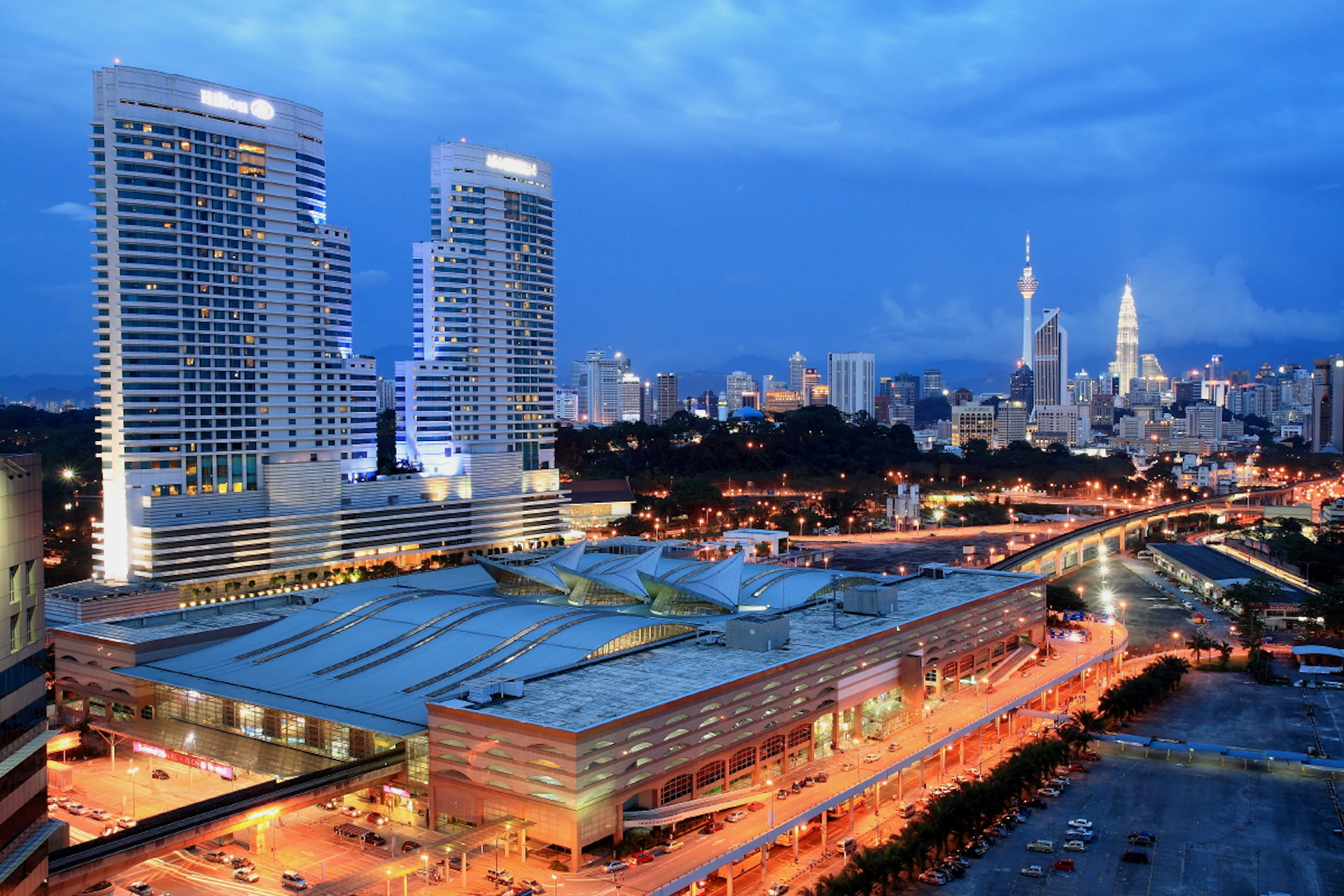 A cityscape at dusk with modern skyscrapers, a lit-up building complex in the foreground, and a prominent tower in the distance.