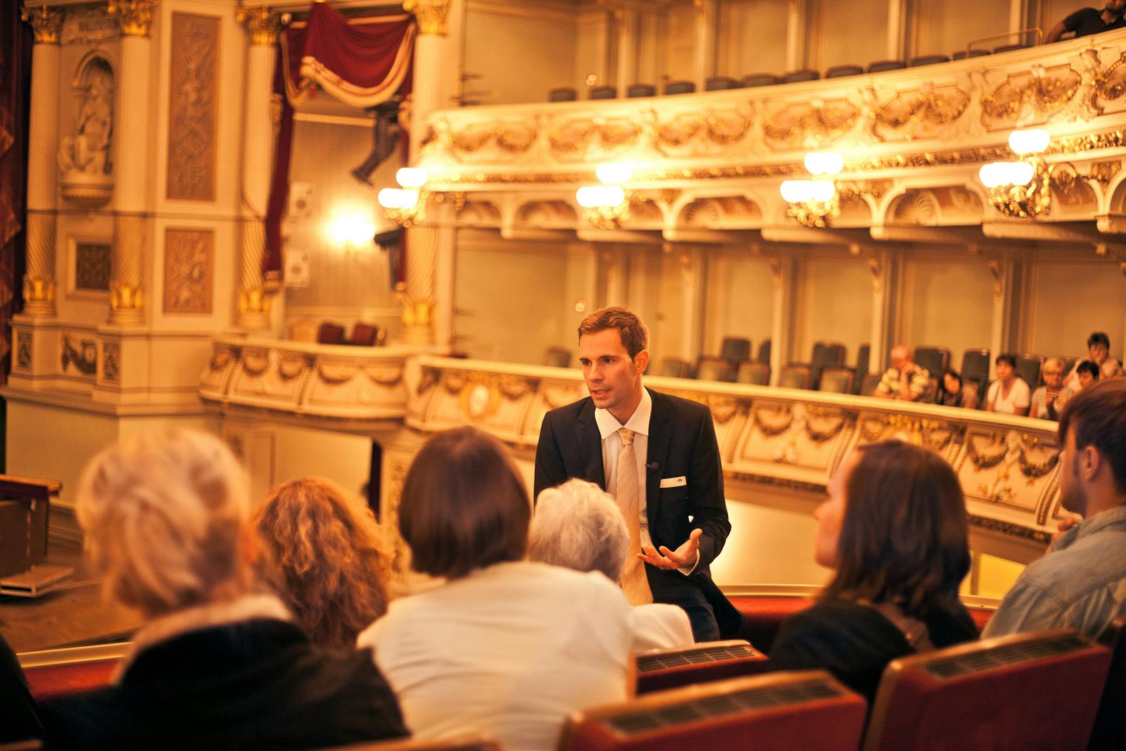 A man in a suit speaks to a small group of people in an ornate theater with elaborate lighting and decor.