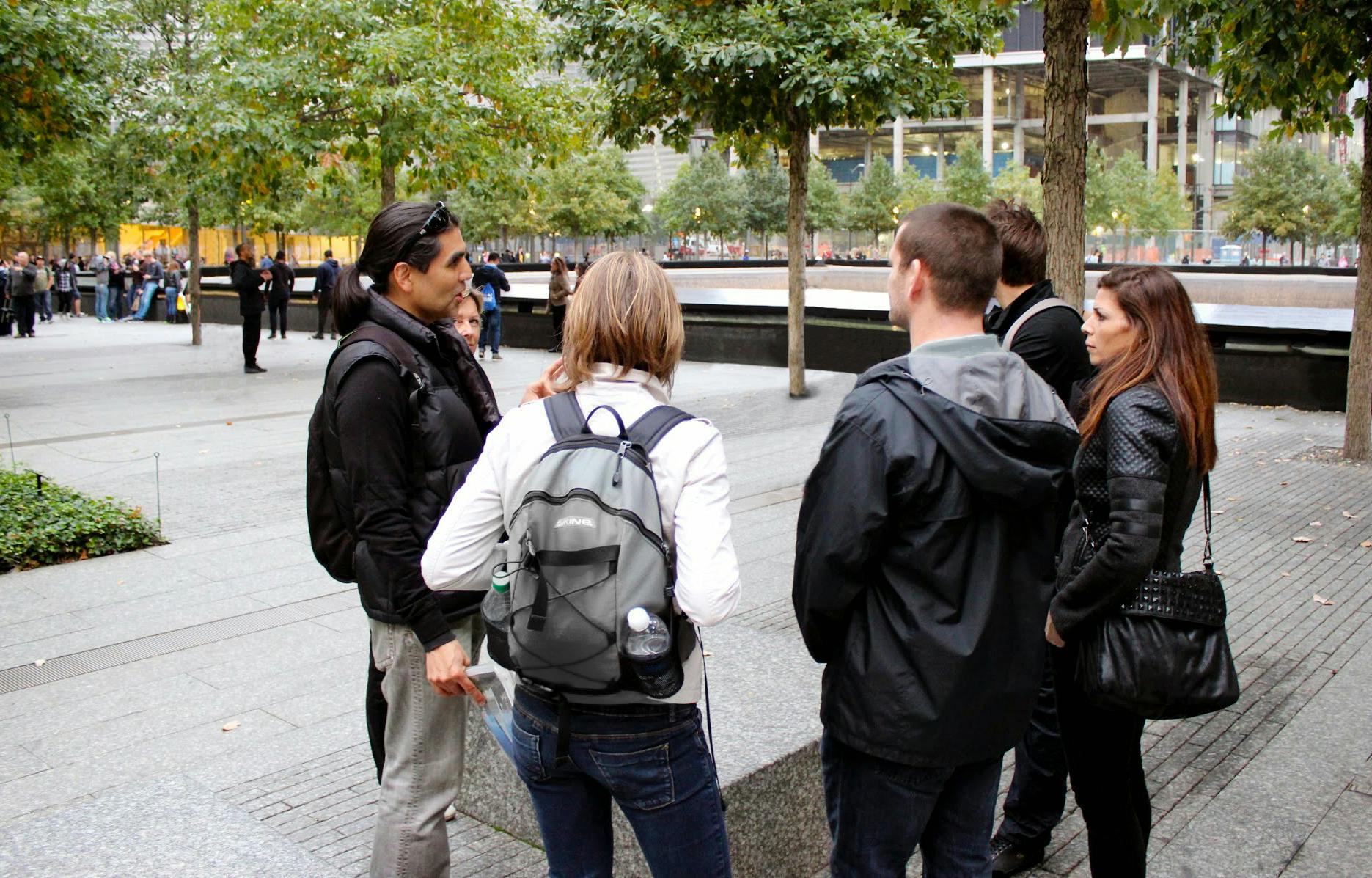 Quatro pessoas estão junto a uma piscina memorial, rodeadas por árvores e estruturas urbanas. Uma pessoa tem uma mochila.