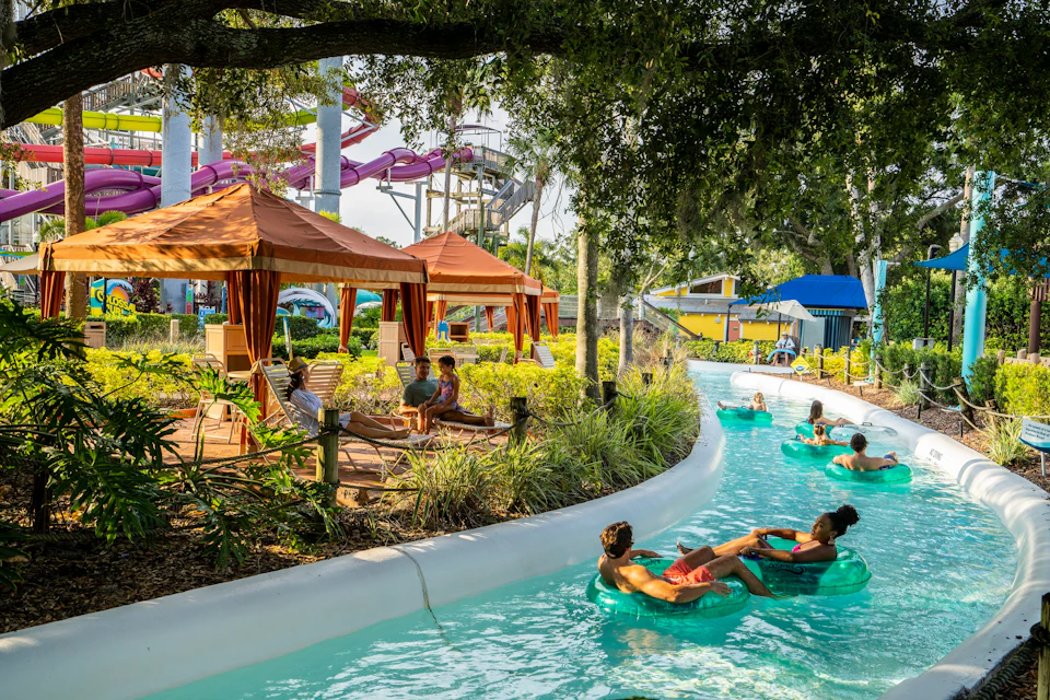People relax on inflatable tubes in a lazy river near cabanas and waterslides in a lush, shaded water park.