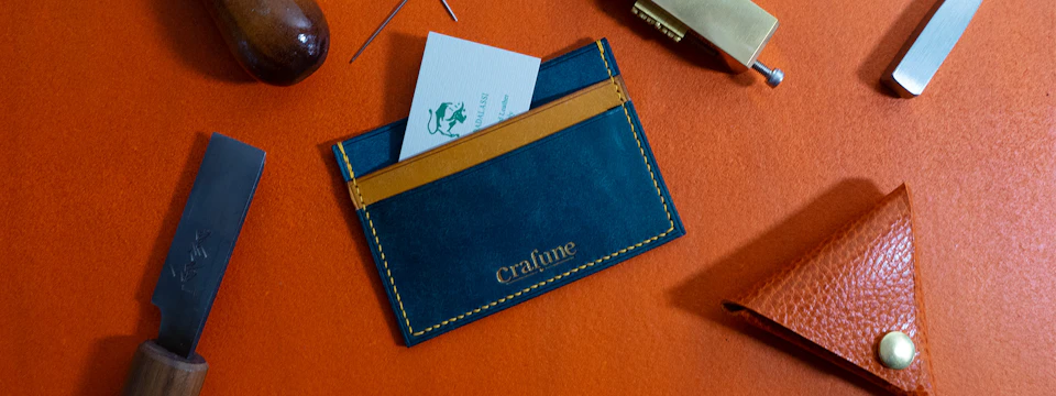 A blue and tan leather cardholder with "Crafune" embossed, holding a business card, placed on an orange surface.